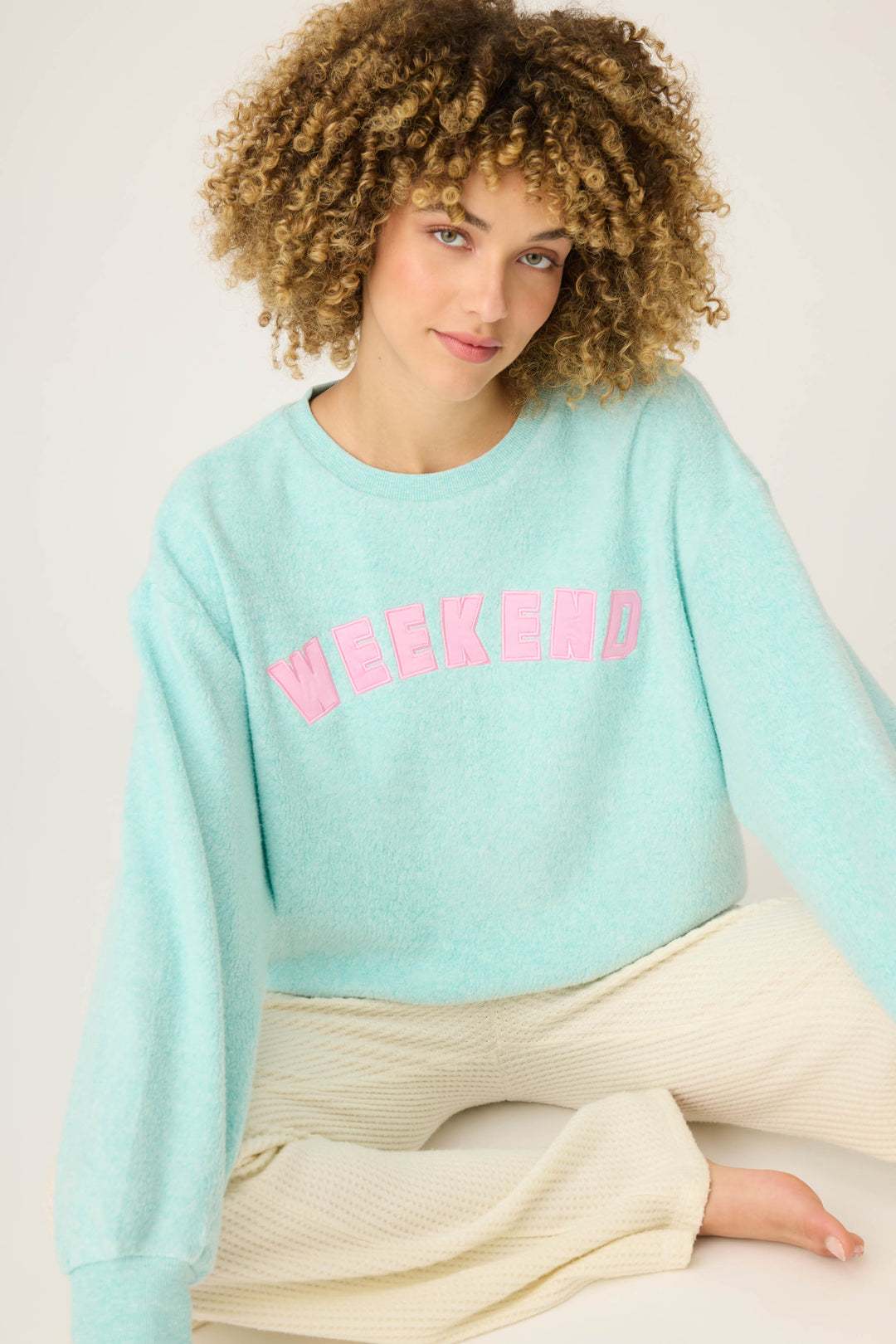 A person with curly hair sits on the floor against a plain light background, wearing P.J. Salvages Secret Garden Weekend Graphic Sweatshirt in light blue and cream-colored textured pants.