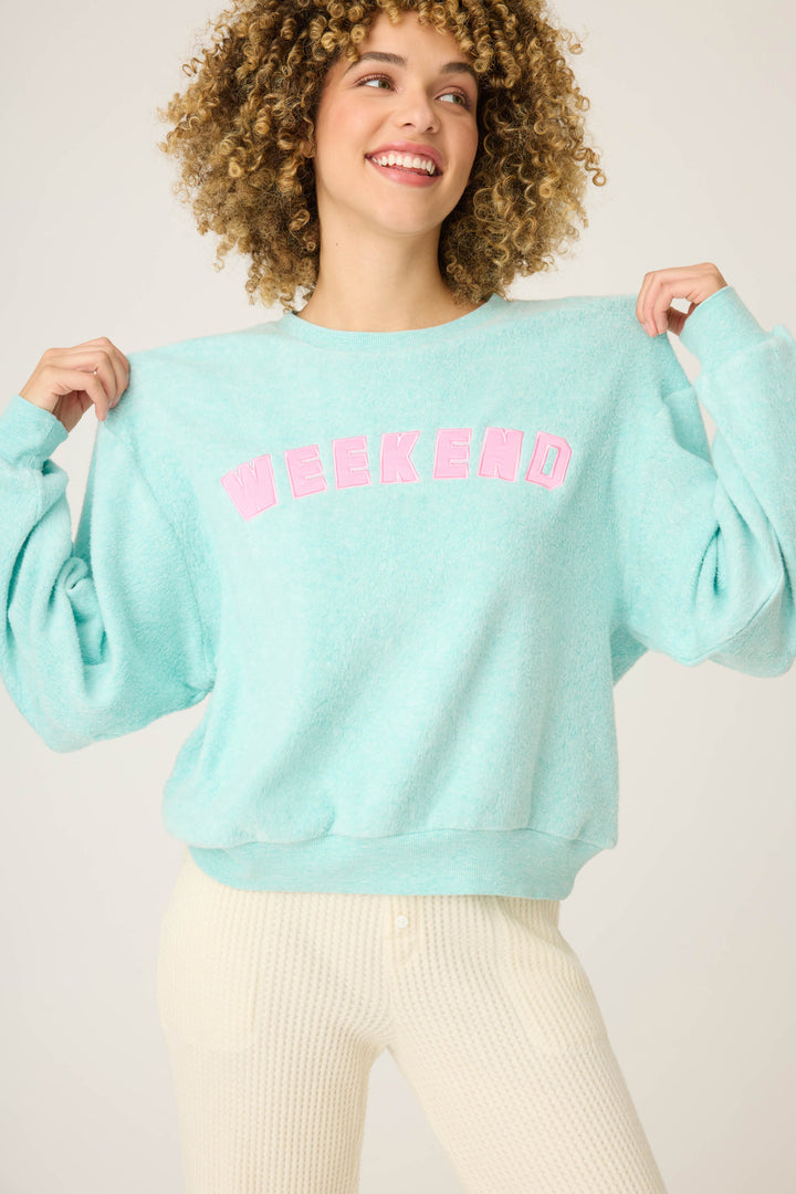 A person with curly hair smiles, holding up the Secret Garden Weekend Graphic Sweatshirt by P.J. Salvage—light blue with WEEKEND in pink—and pairs it with light-colored pants.