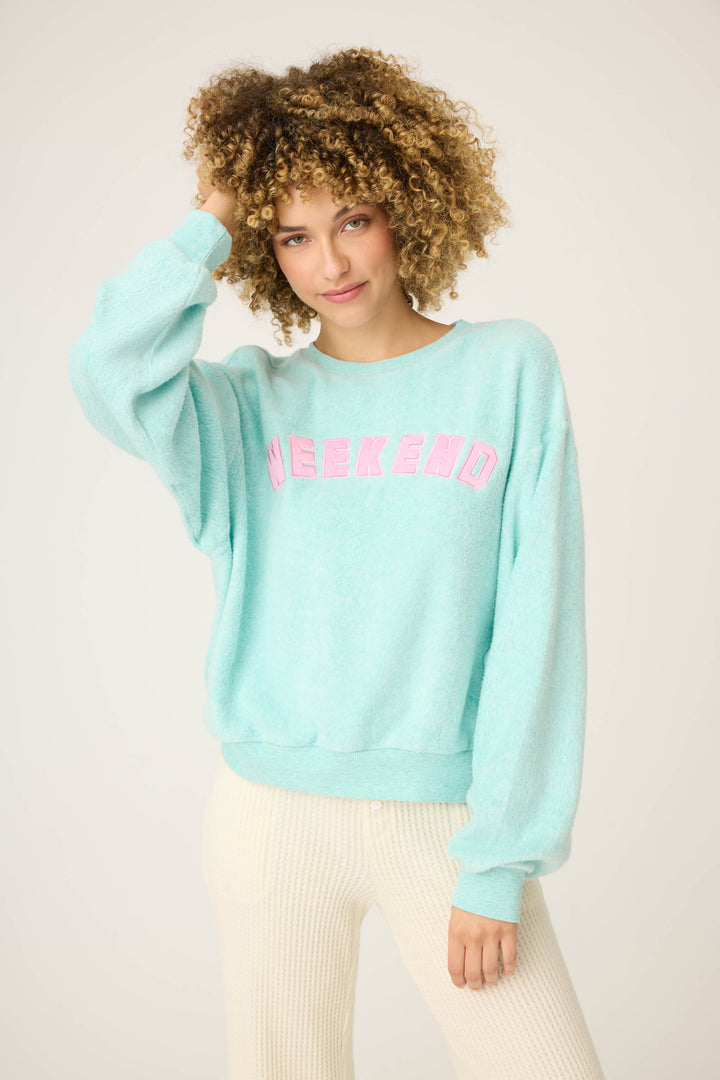 A person with curly hair wears the P.J. Salvage Secret Garden Weekend Graphic Sweatshirt in light blue with WEEKEND in pink and cream-colored pants, standing against a plain background, one hand touching their hair.