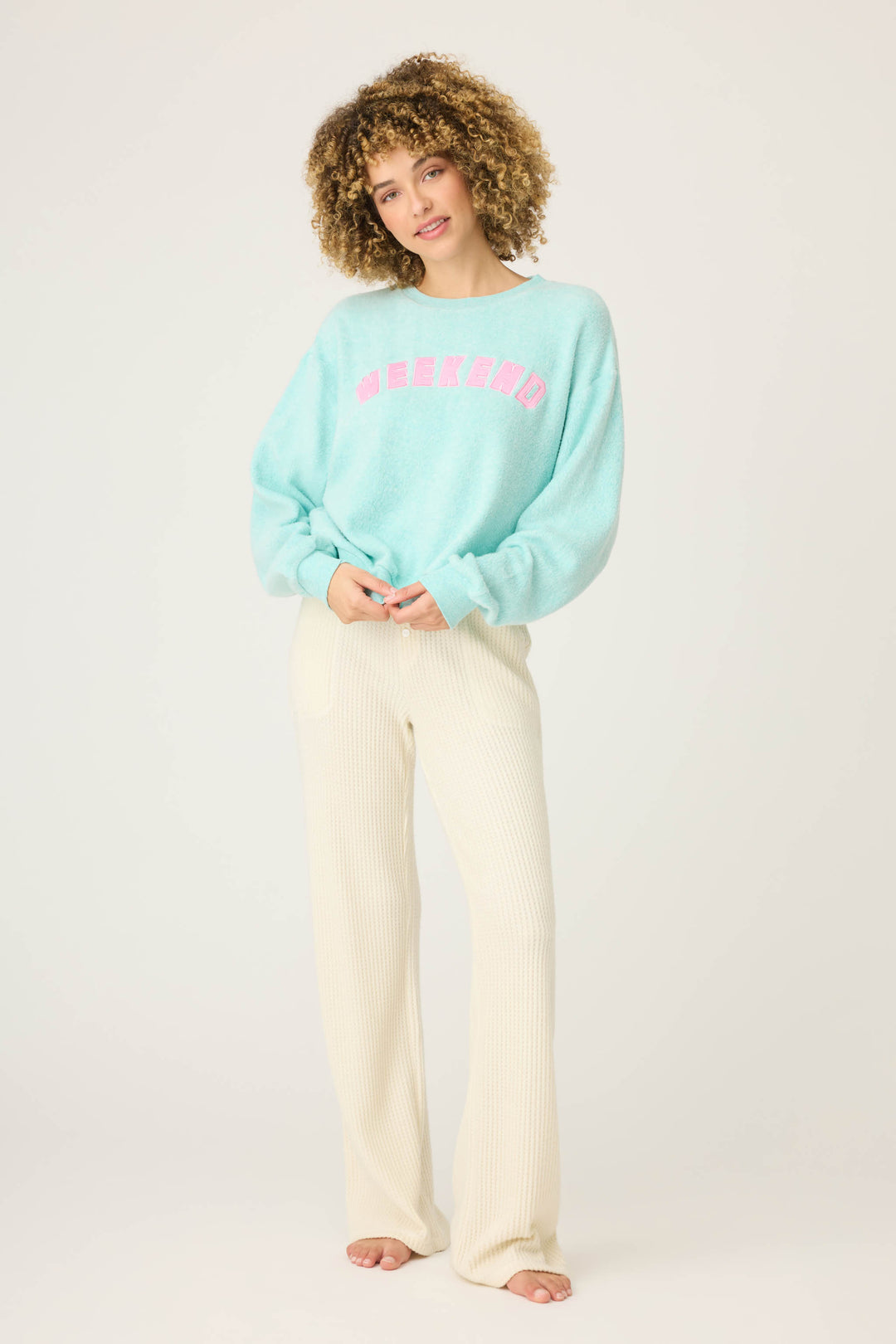 A person with curly hair poses barefoot against a plain backdrop, wearing cream ribbed pants and the P.J. Salvage Secret Garden Weekend Graphic Sweatshirt—a loose light blue top with WEEKEND in pink across the chest.