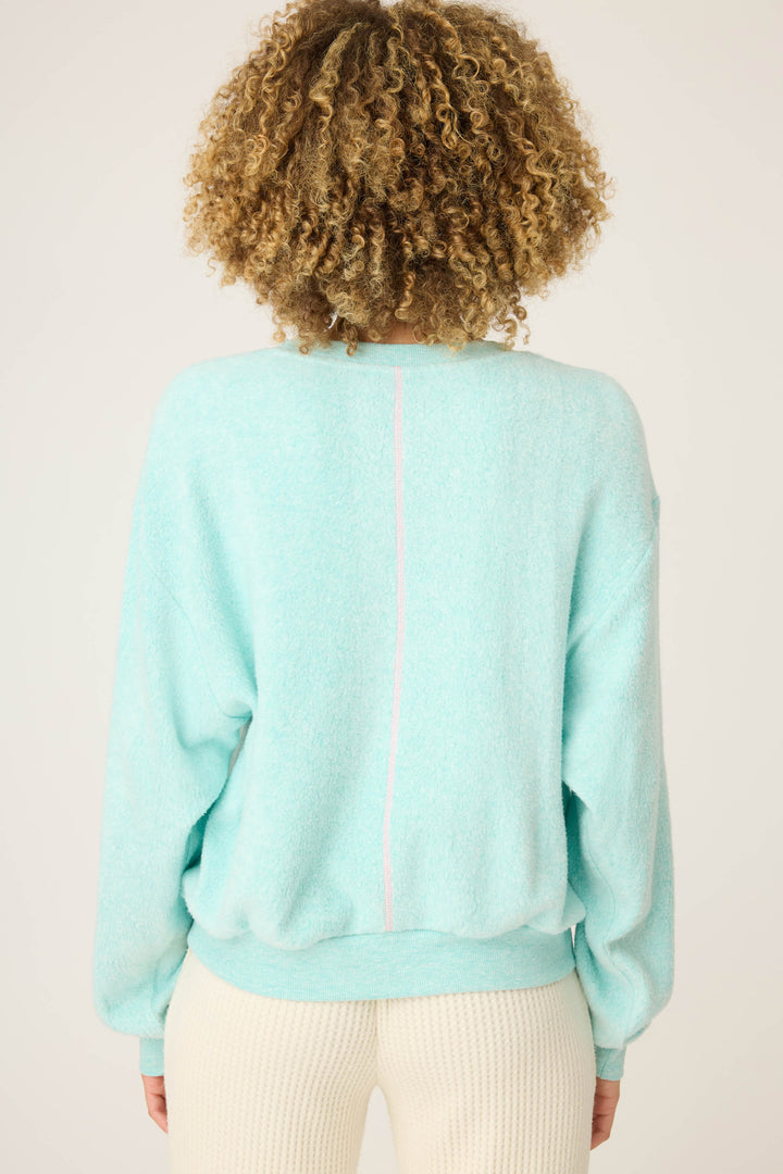 A person with curly hair is seen from behind wearing the P.J. Salvage Secret Garden Weekend Graphic Sweatshirt—a loose, light blue top with a subtle pink center line—paired with cream textured pants against a plain, light background.