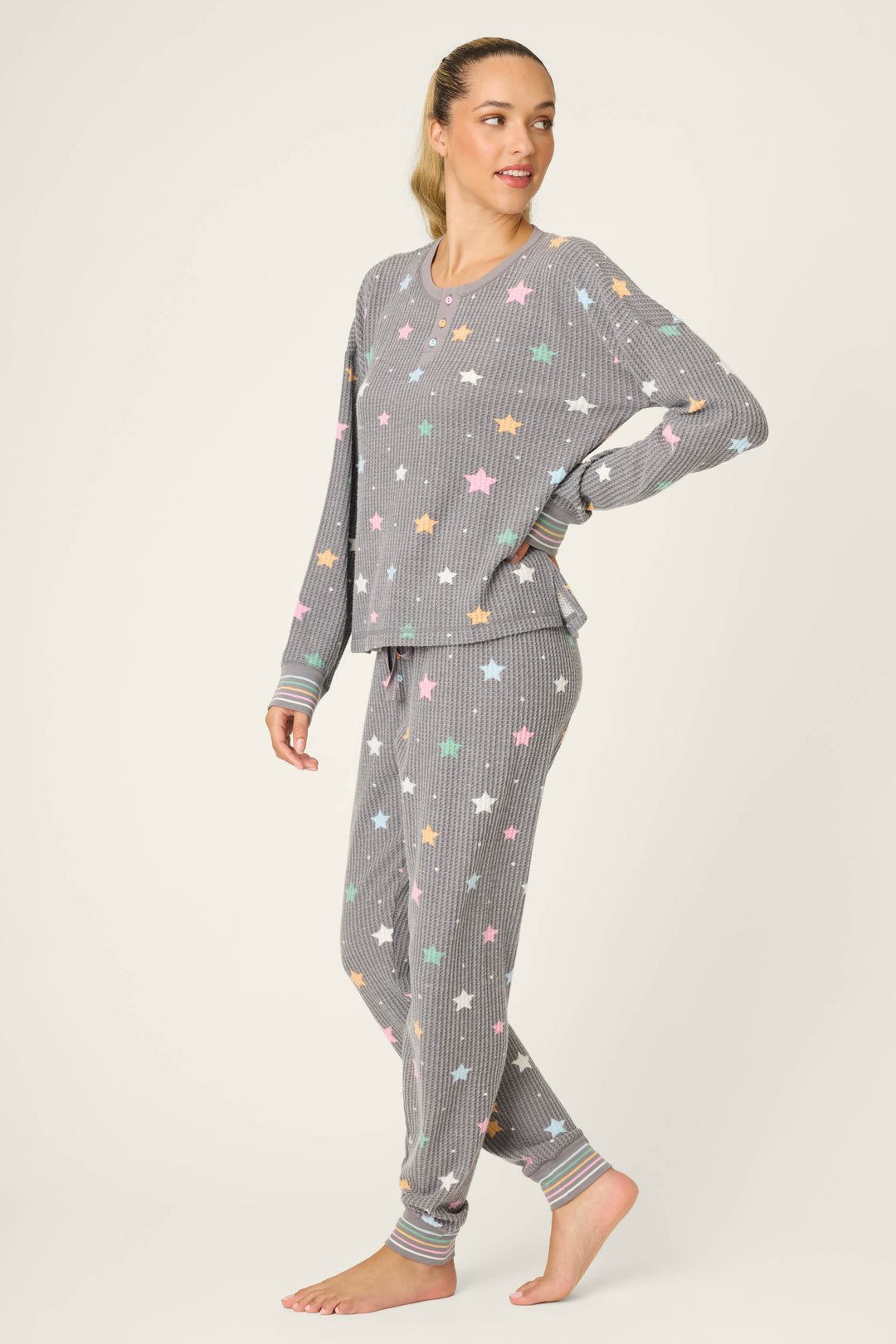 A woman with her hair in a ponytail stands barefoot, wearing the P.J. Salvage Ski Jammie Classics Star Print PJ Set, a matching long-sleeve pajama set with grey and white stripes and multicolored stars.