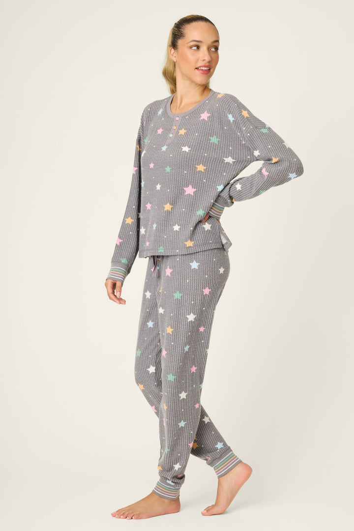 A woman with her hair in a ponytail stands barefoot, wearing the P.J. Salvage Ski Jammie Classics Star Print PJ Set, a matching long-sleeve pajama set with grey and white stripes and multicolored stars.