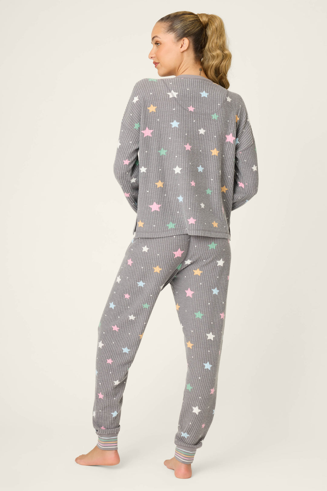 A person stands barefoot facing away, wearing the P.J. Salvage Ski Jammie Classics Star Print PJ Set—gray ribbed pajamas with colorful stars and striped cuffs—against a plain light background.
