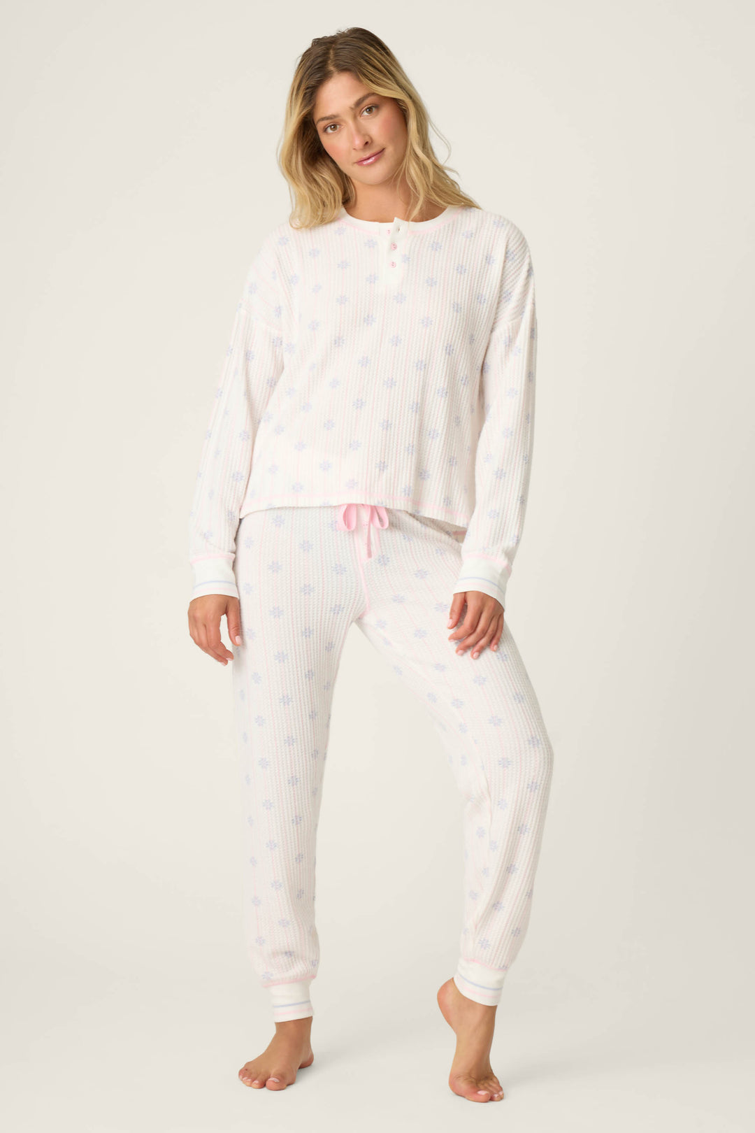 A woman stands barefoot against a plain light background wearing the P.J. Salvage Ski Jammie Classics Snowflake Print PJ Set, a long-sleeved, light pajama set with a subtle snowflake pattern and pink drawstring at the waist.