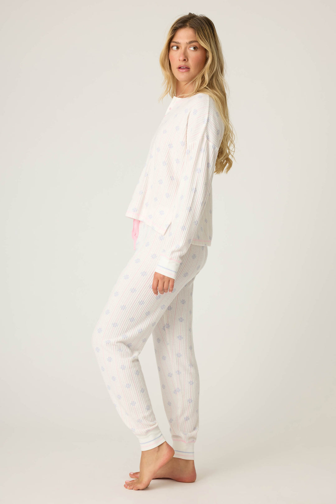A woman stands barefoot against a plain background, wearing the P.J. Salvage Ski Jammie Classics Snowflake Print PJ Set with light blue patterns. She faces sideways, head turned slightly toward the camera, her long hair down.