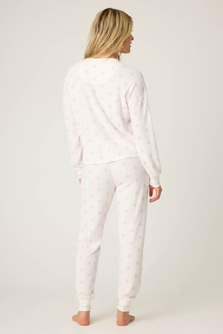 A person stands barefoot facing away, wearing the P.J. Salvage Ski Jammie Classics Snowflake Print PJ Set: a long-sleeve white pajama with subtle blue snowflakes and ribbed cuffs, set against a plain, light background.