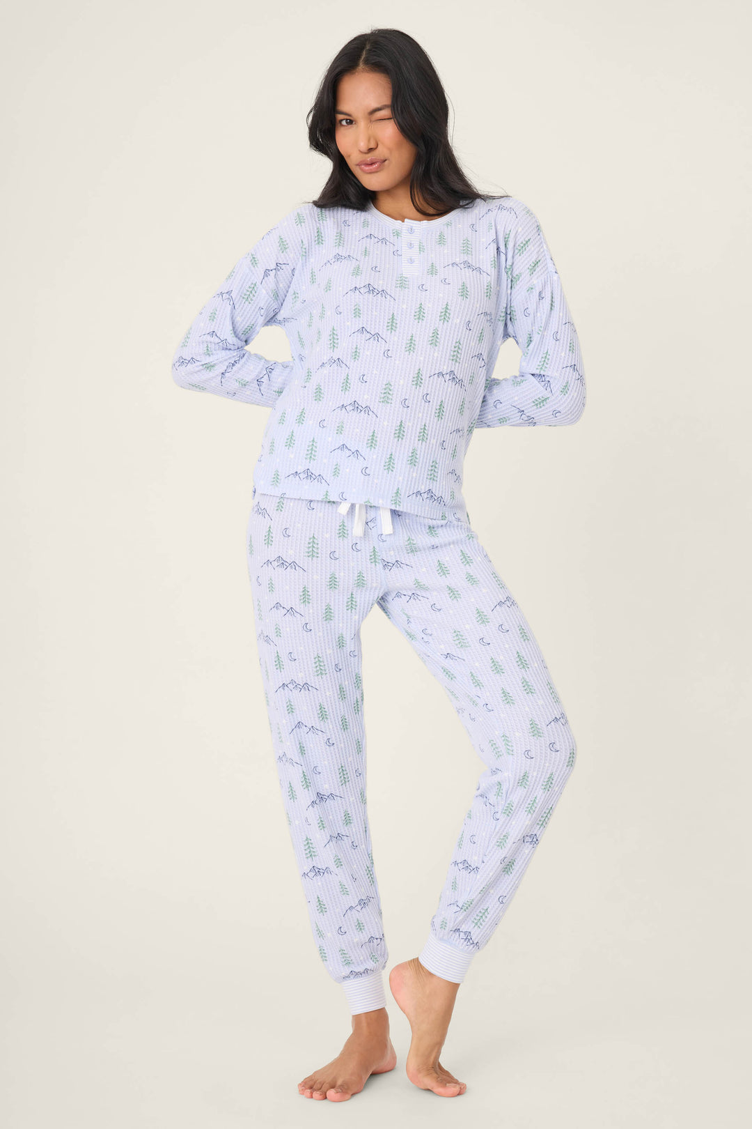 A person with long dark hair stands barefoot on a plain background, winking and posing in the P.J. Salvage Ski Jammie Classics Winter Tree Print PJ Set.