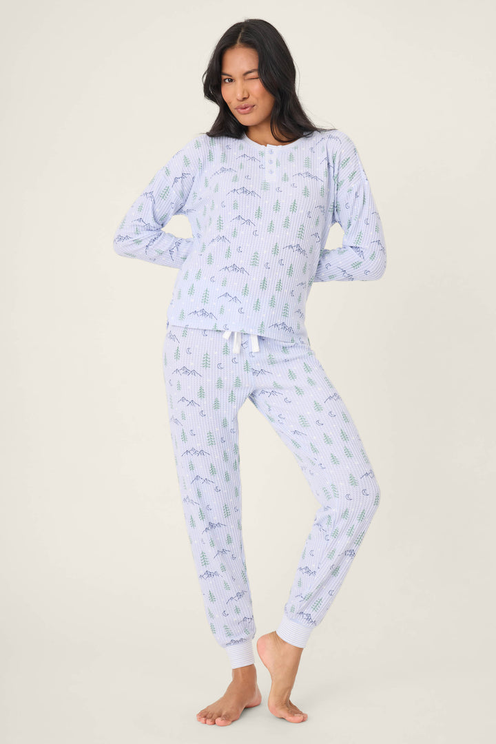 A person with long dark hair stands barefoot on a plain background, winking and posing in the P.J. Salvage Ski Jammie Classics Winter Tree Print PJ Set.