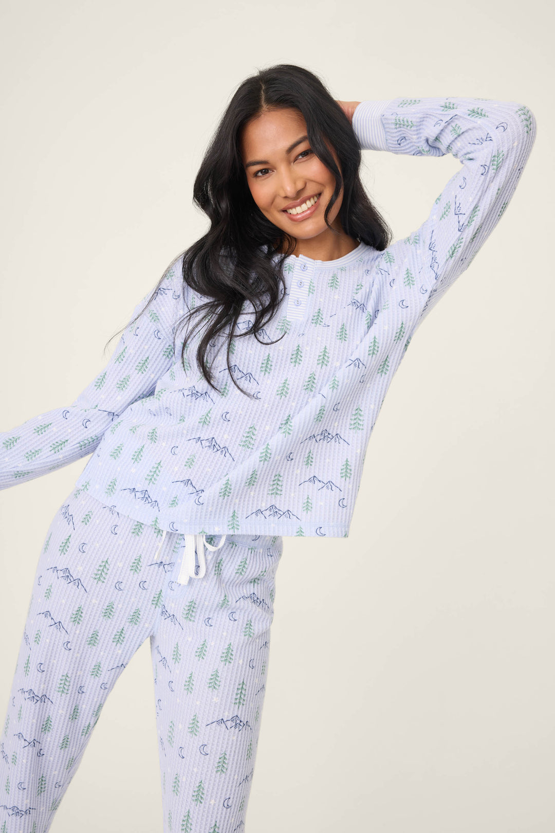 A woman with long black hair smiles and poses with one hand behind her head, wearing the P.J. Salvage Ski Jammie Classics Winter Tree Print PJ Set in light blue against a neutral background.