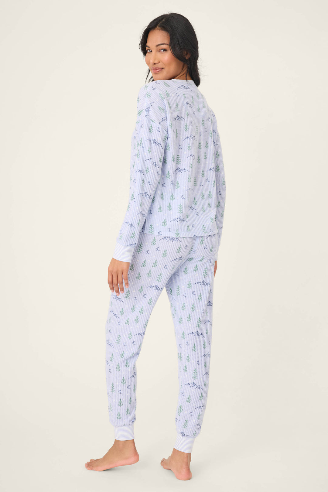 A woman stands barefoot, turned slightly away, wearing the P.J. Salvage Ski Jammie Classics Winter Tree Print PJ Set—light blue pajama pants and a long-sleeve top with green and purple designs—against an off-white background.