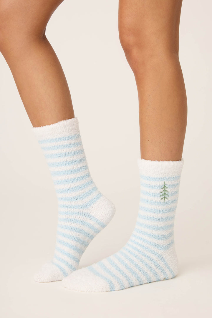 A person wears P.J. Salvage Striped Christmas Tree Fuzzy Crew Socks featuring cozy blue and white stripes and a green tree embroidered near the top. The background is plain and light-colored.