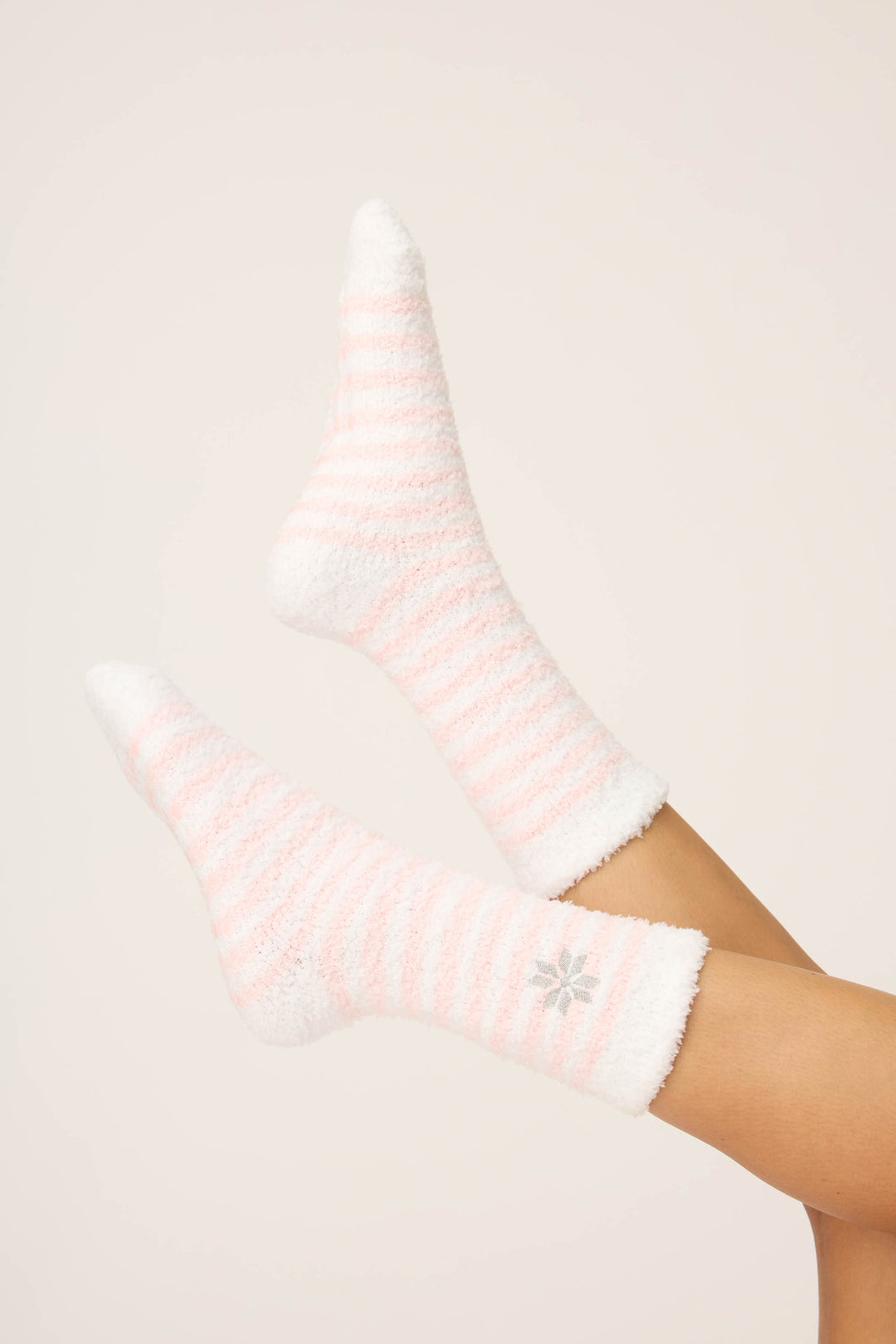 A person’s legs are raised, wearing P.J. Salvage Striped Snowflake Fuzzy Crew Socks in white with light pink stripes and a gray snowflake design, set against a plain light background.