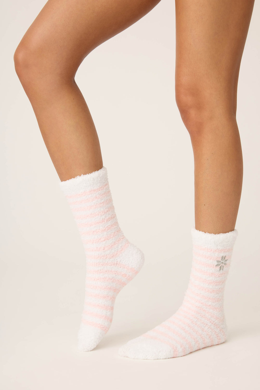 A person wears P.J. Salvage Striped Snowflake Fuzzy Crew Socks—white with light pink stripes and a gray snowflake near the top—standing against a plain, light background with one leg slightly bent.