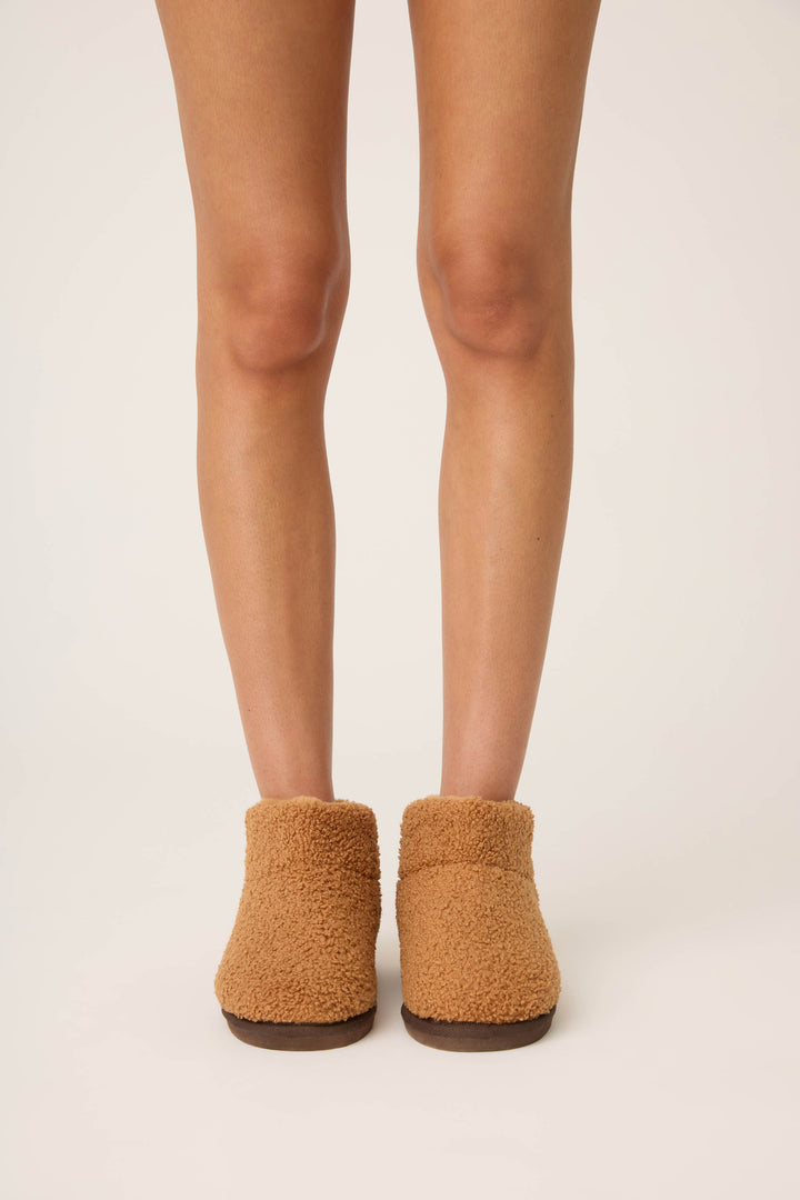 A person stands on a light surface, shown from mid-thigh down, wearing P.J. Salvage Teddy Bear Sherpa Booties—tan, fuzzy, closed-toe plush slippers.