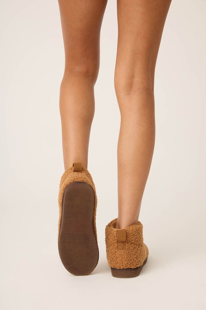 A person is shown from the knees down, standing on a light surface and wearing P.J. Salvage Teddy Bear Sherpa Booties in tan—one foot flat, the other lifted to reveal the plush slipper’s sole.