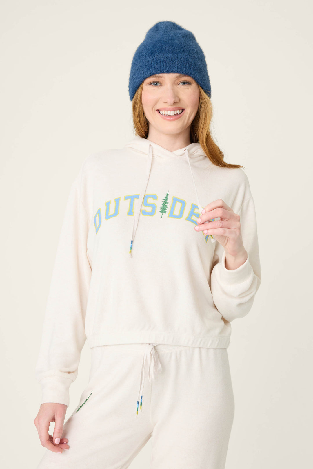 A person wears The Outsider Fleece Graphic Hoodie by P.J. Salvage, featuring "OUTSIDER" and a tree graphic, along with a blue knit beanie. They are smiling and standing against a plain light background.