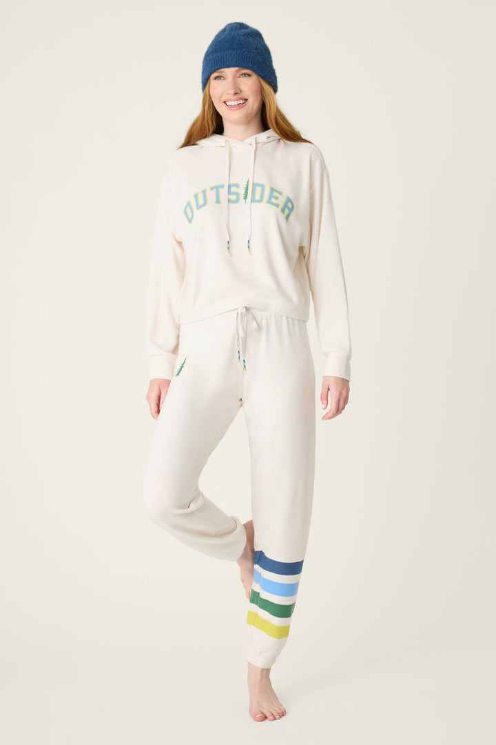 A woman stands barefoot on one leg wearing the P.J. Salvage The Outsider Fleece Graphic Hoodie with "OUTSIDER" in green and blue, a blue beanie, and cream sweatpants with green, blue, and yellow stripes by her left ankle.