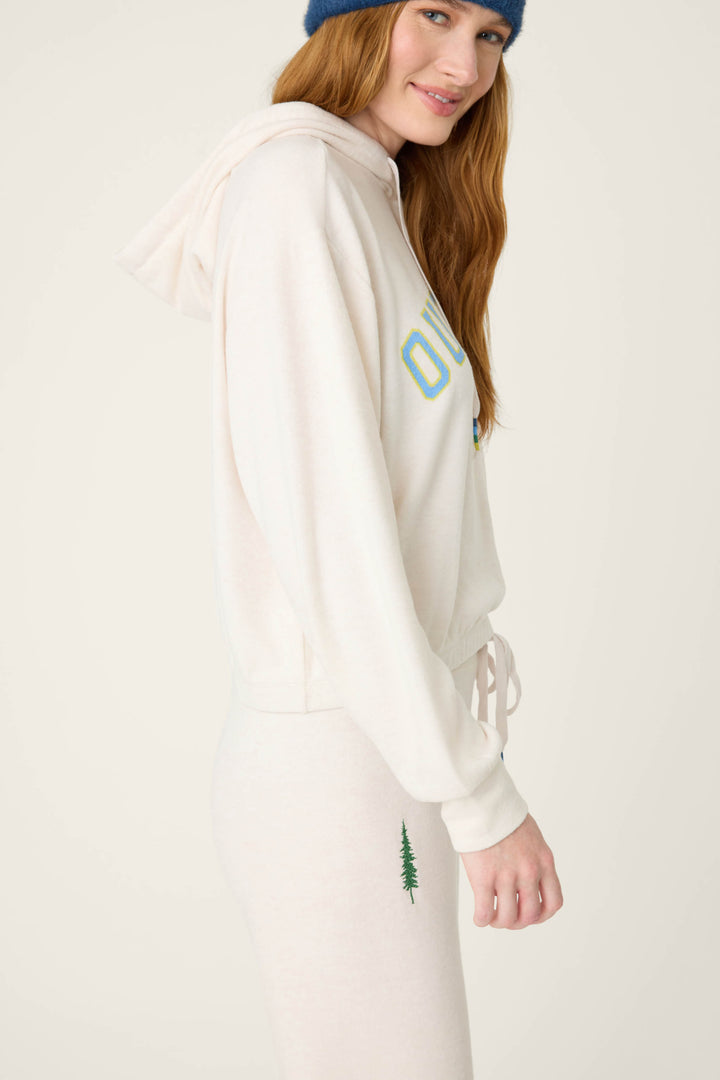 A person with long red hair stands sideways, smiling slightly. They wear the P.J. Salvage The Outsider Fleece Graphic Hoodie in cream and matching sweatpants, featuring a small green tree embroidery, plus a blue knit hat.