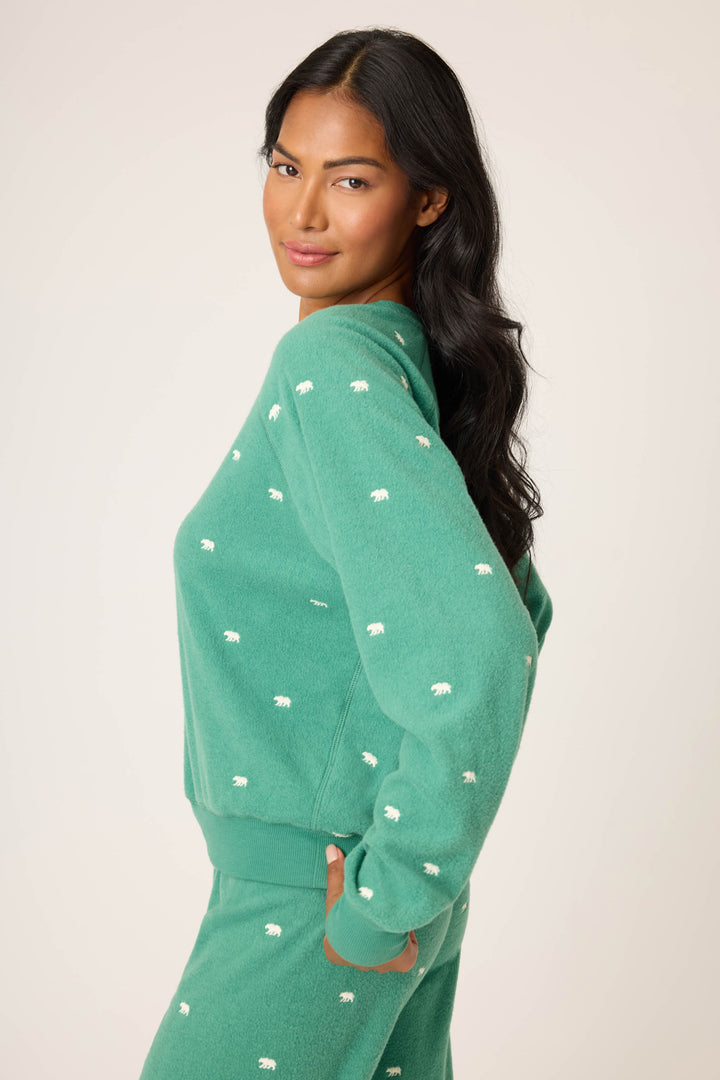 A person with long, wavy black hair stands sideways against a light background, wearing the P.J. Salvage The Outsider Embroidered Bear Fleece Sweatshirt and looking at the camera with one hand on their hip.