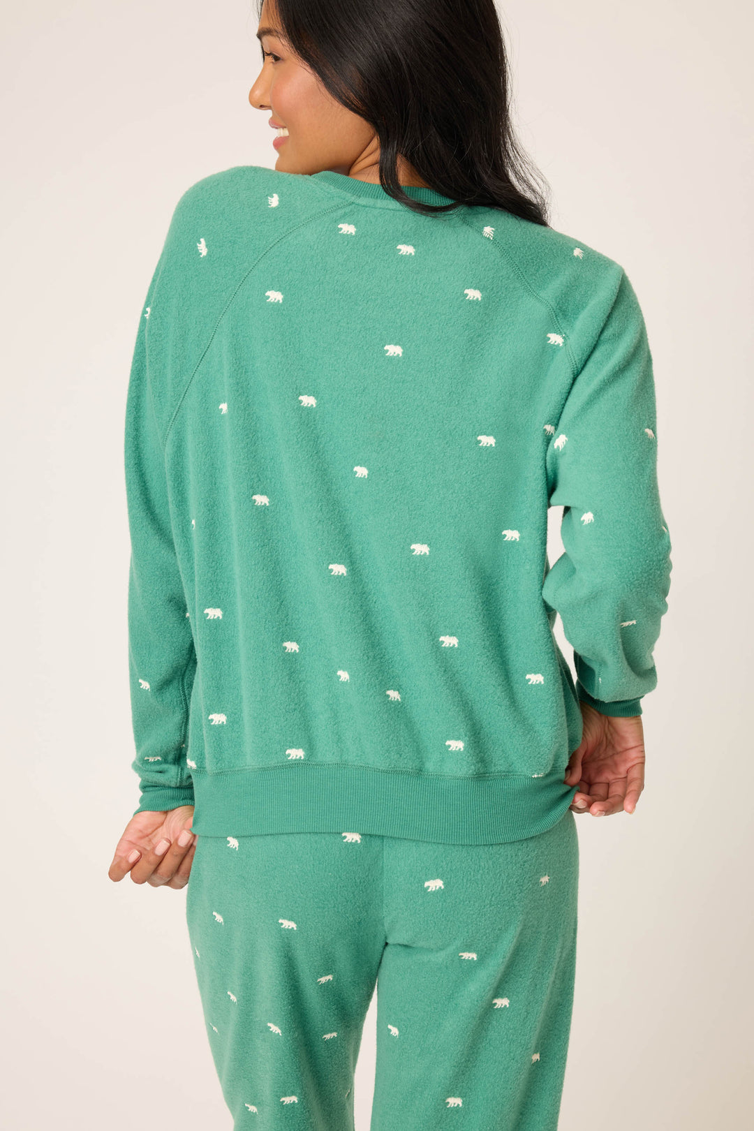 A person with long dark hair is seen from the back adjusting the hem of a green P.J. Salvage The Outsider Embroidered Bear Fleece Sweatshirt featuring a small white bear pattern and matching pants.