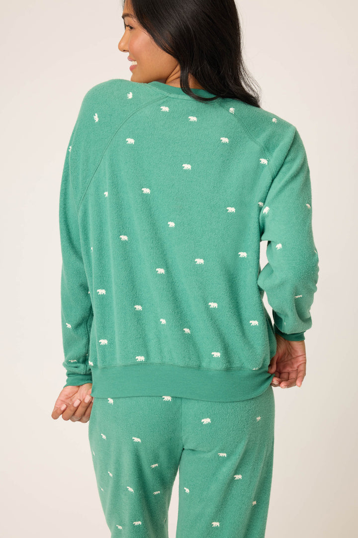 A person with long dark hair is seen from the back adjusting the hem of a green P.J. Salvage The Outsider Embroidered Bear Fleece Sweatshirt featuring a small white bear pattern and matching pants.