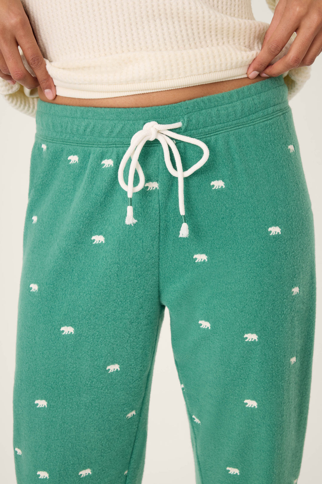 A person wears P.J. Salvage's The Outsider Embroidered Bear Fleece Banded Pants—green with white bear prints and a white drawstring—paired with a cream-colored ribbed top. Image is cropped from waist to mid-thigh.