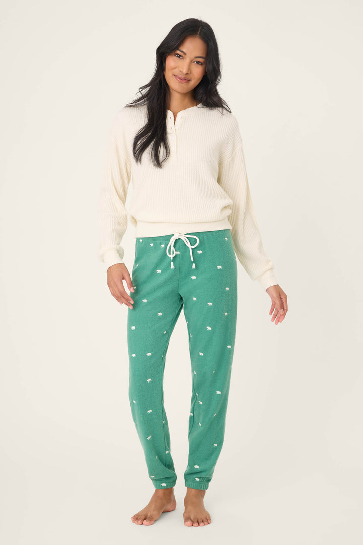 A person stands barefoot against a plain background, wearing a cream long-sleeve knit top and The Outsider Embroidered Bear Fleece Banded Pants by P.J. Salvage. They have long dark hair and a relaxed expression.
