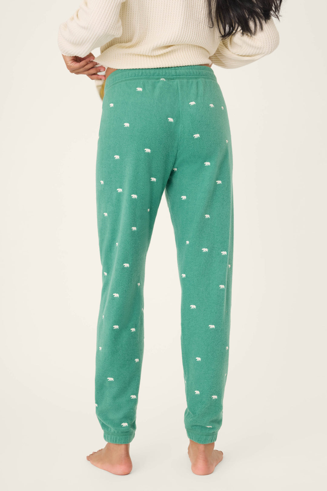 A person stands barefoot facing away, wearing P.J. Salvage's The Outsider Embroidered Bear Fleece Banded Pants in green with a small white bear print and a cream sweater, against a plain light background.