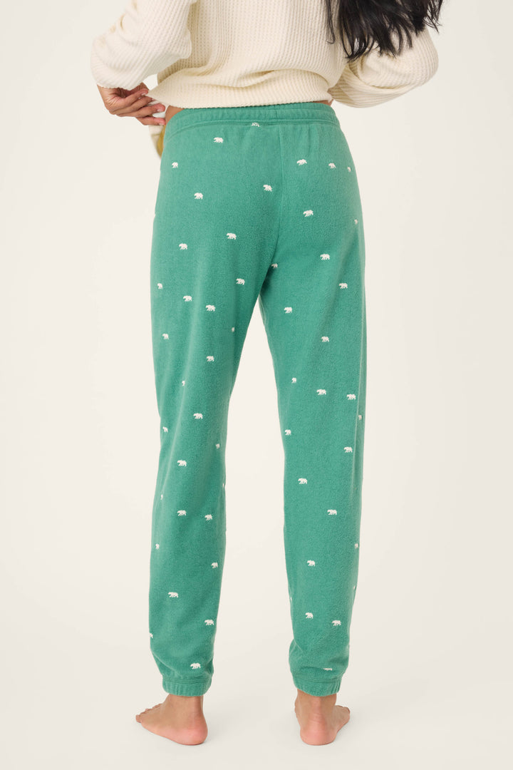 A person stands barefoot facing away, wearing P.J. Salvage's The Outsider Embroidered Bear Fleece Banded Pants in green with a small white bear print and a cream sweater, against a plain light background.