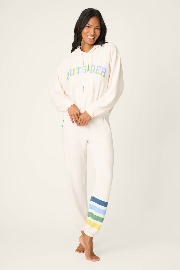 A woman smiles barefoot, wearing P.J. Salvage’s The Outsider Graphic Jogger Lounge Set—featuring a cream hoodie with "OUTSIDER" text and matching joggers detailed with blue, green, and yellow stripes at the left ankle.