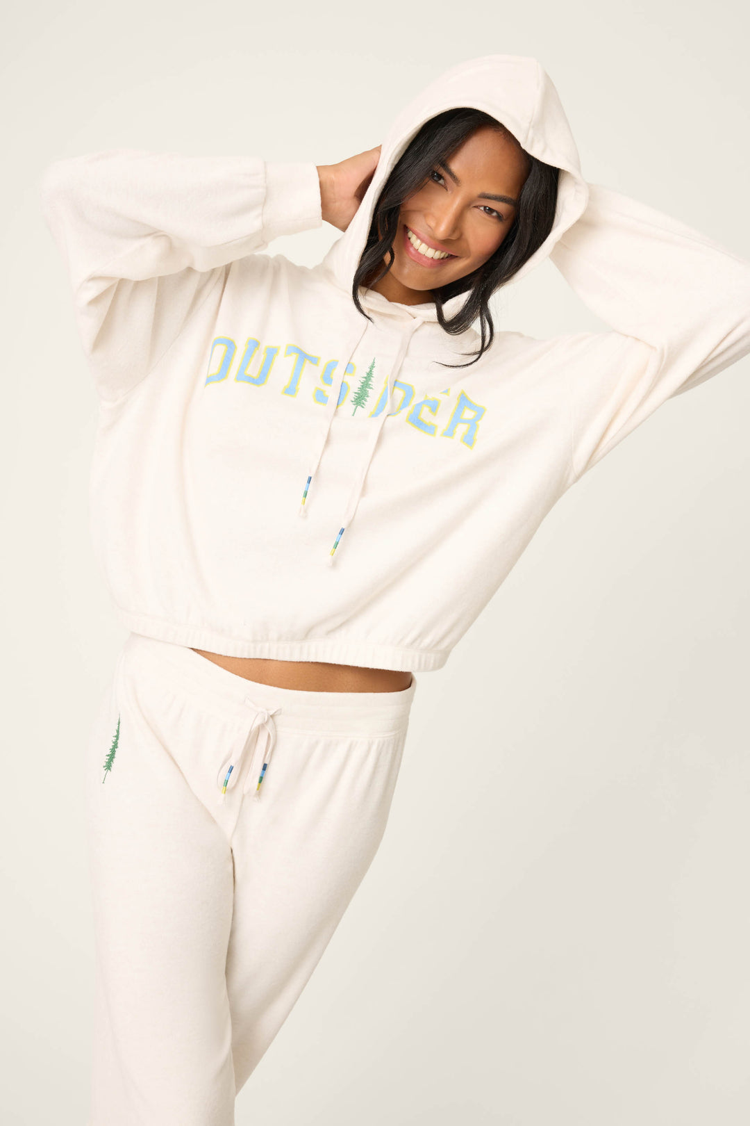 A person smiles against a plain background, holding the hood of the P.J. Salvage The Outsider Graphic Jogger Lounge Set—featuring a cream-colored hoodie with "OUTSIDER" on it and matching sweatpants.