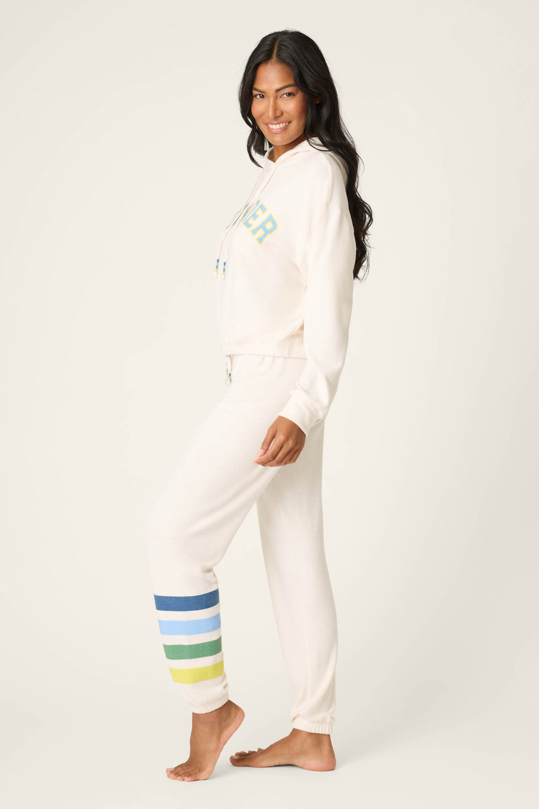 A woman smiles while standing barefoot on one leg, wearing the P.J. Salvage The Outsider Graphic Jogger Lounge Set in cream with multicolored stripes, posing sideways against a plain light background.