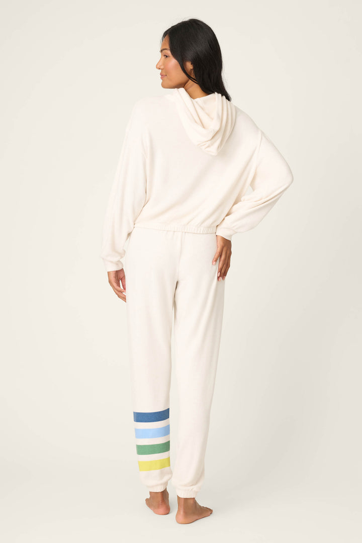 A person stands barefoot facing away, wearing the P.J. Salvage "The Outsider Graphic Jogger Lounge Set"—a loose-fitting, cream-colored hooded sweatshirt and matching sweatpants with blue, green, and yellow stripes on one ankle.
