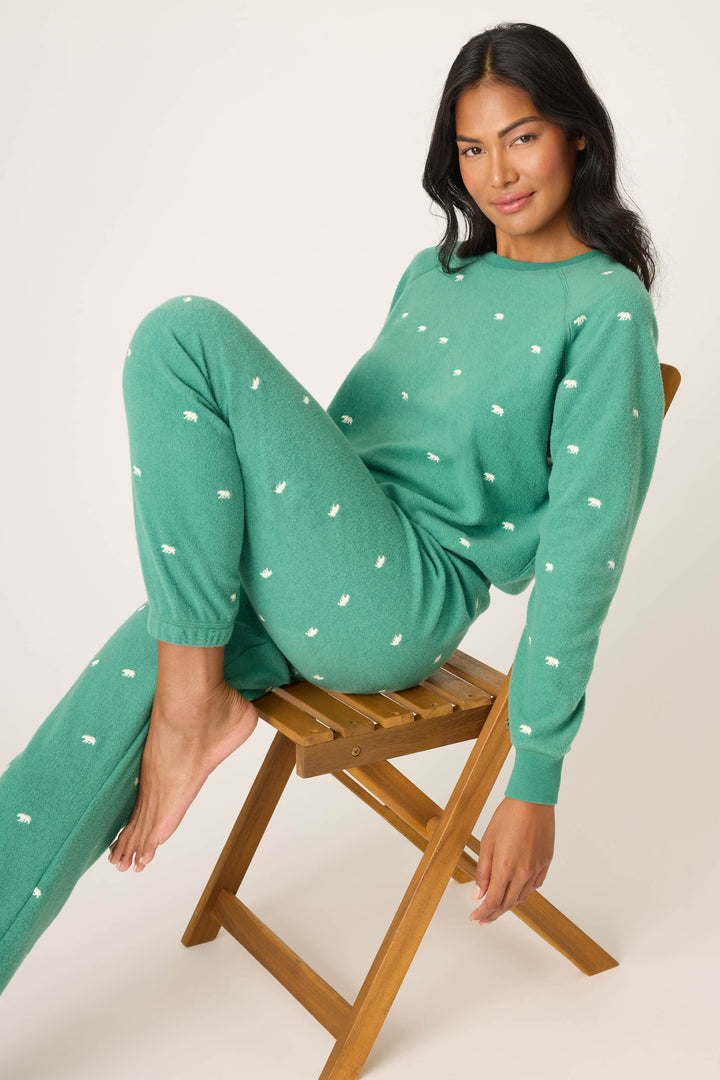 A woman with long dark hair sits on a wooden folding chair, barefoot and relaxed, wearing P.J. Salvage's The Outsider Embroidered Bear Fleece Lounge Set in green with white embroidered details.