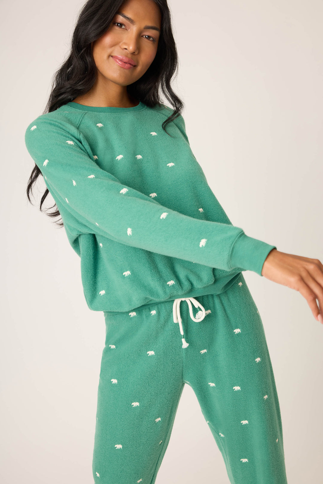 A person models the P.J. Salvage "The Outsider Embroidered Bear Fleece Lounge Set," featuring a matching green long-sleeve top and drawstring pants with small white bear designs, standing in a relaxed pose against a neutral background.