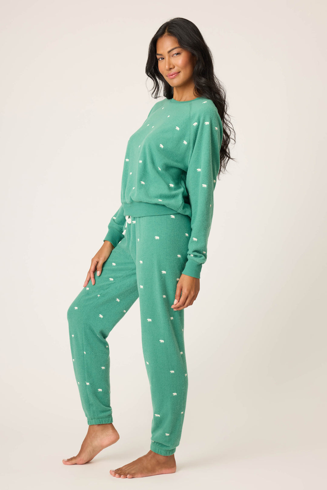 A person stands barefoot on one foot, wearing the P.J. Salvage The Outsider Embroidered Bear Fleece Lounge Set—a matching green pajama set with white dots, featuring a long-sleeve top and drawstring pants—against a plain, light background.