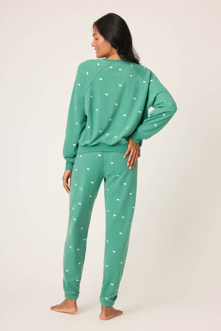 A person stands barefoot facing away in the P.J. Salvage "The Outsider Embroidered Bear Fleece Lounge Set"—a green matching sweatshirt and sweatpants, both with small white polka dots. One hand is on their hip, in front of a plain light background.