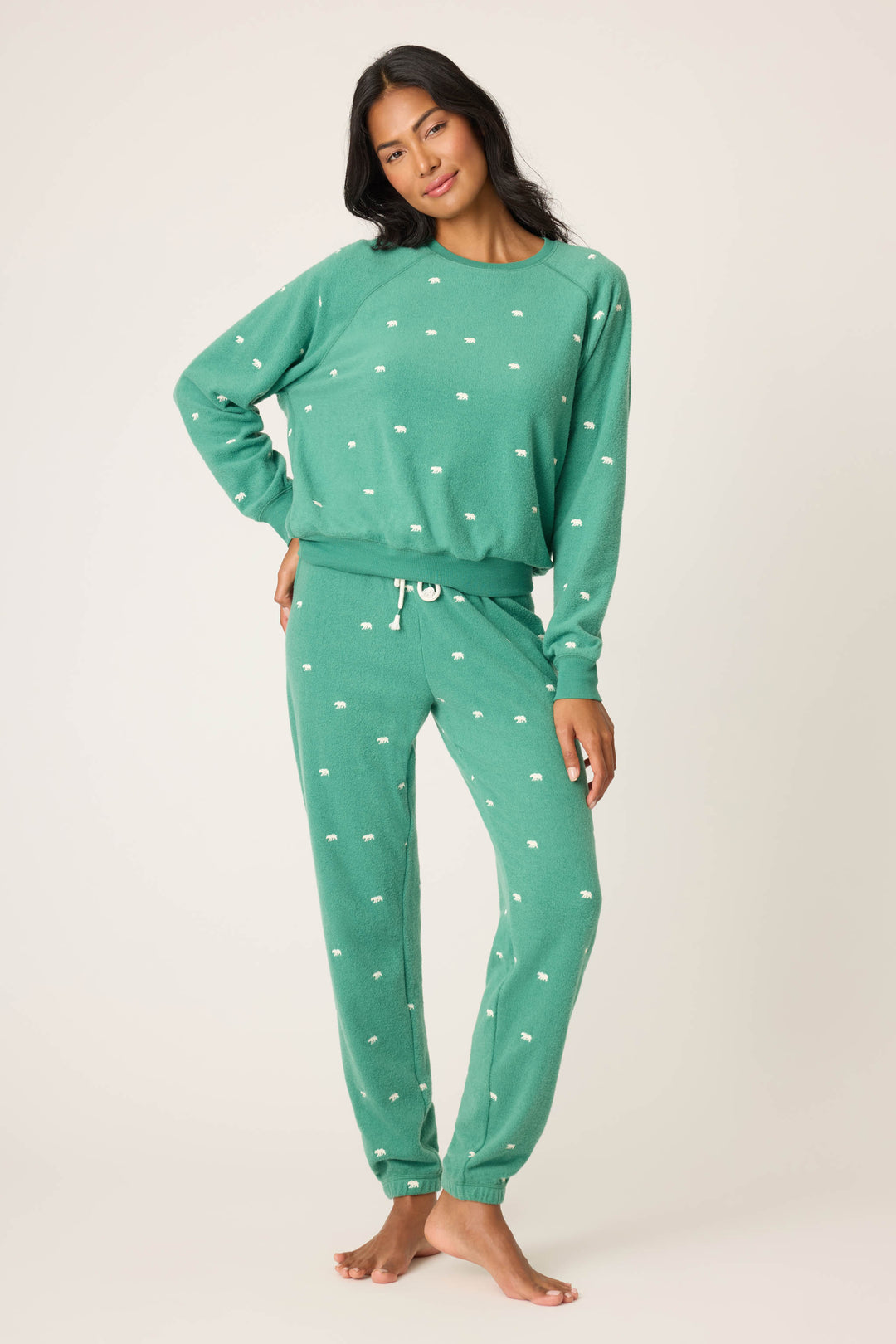 A woman stands barefoot against a plain background, wearing the P.J. Salvage The Outsider Embroidered Bear Fleece Lounge Set—matching green long-sleeve sweatshirt and jogger pants with small white dot patterns—as she faces the camera, one hand on her hip.