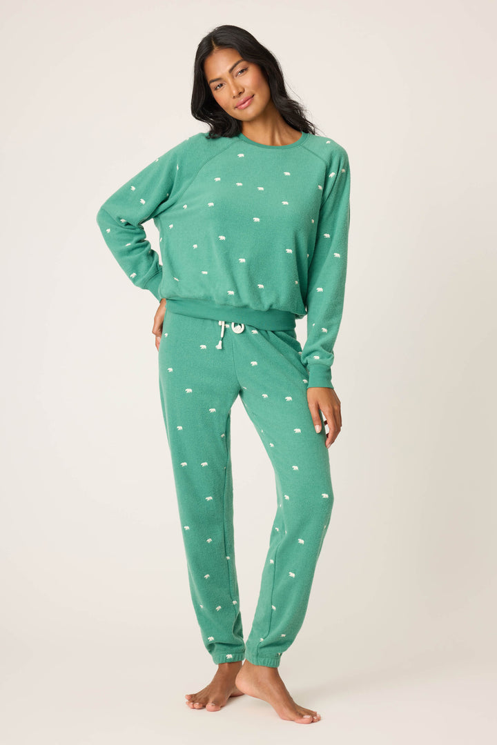 A woman stands barefoot against a plain background, wearing the P.J. Salvage The Outsider Embroidered Bear Fleece Lounge Set—matching green long-sleeve sweatshirt and jogger pants with small white dot patterns—as she faces the camera, one hand on her hip.