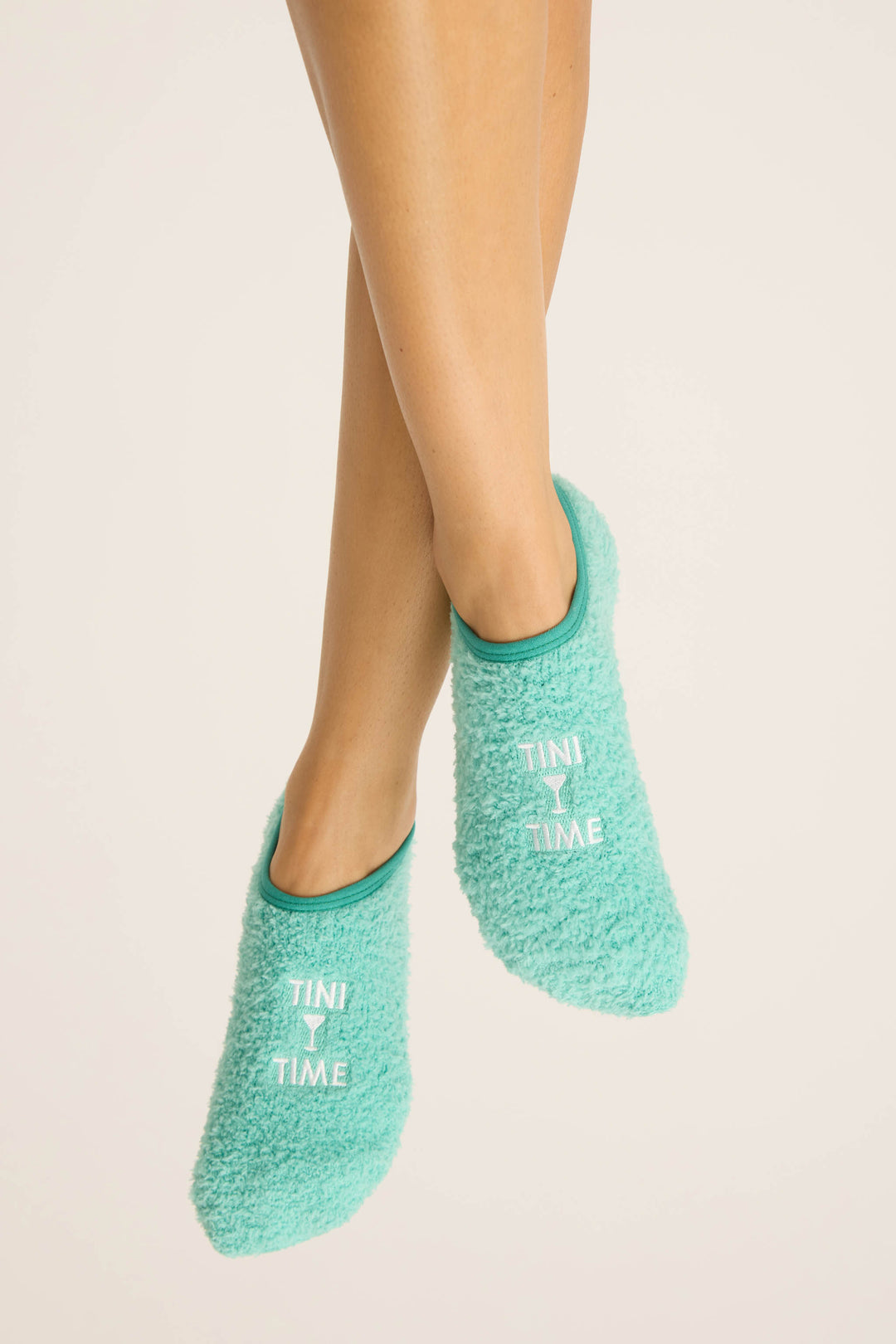 A pair of crossed legs wears P.J. Salvage Tini Time Fuzzy Ankle Socks in teal, featuring “TINI TIME” printed in white on each, set against a plain light background.