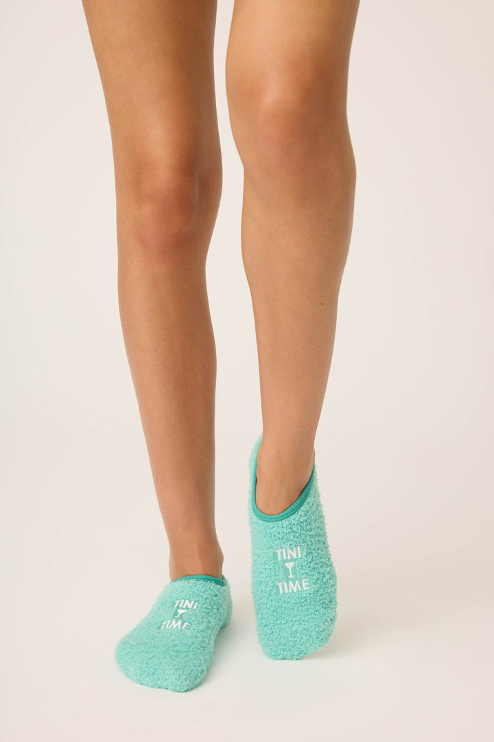 Only the lower legs and feet are visible, showing someone on a light background wearing P.J. Salvage's Tini Time Fuzzy Ankle Socks in turquoise, with "TINY TIME" written in white across the top of each sock.