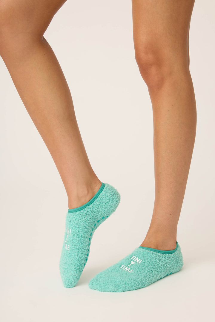 A person stands on a light background showing their lower legs and feet in P.J. Salvage Tini Time Fuzzy Ankle Socks, featuring turquoise fuzz and white text.