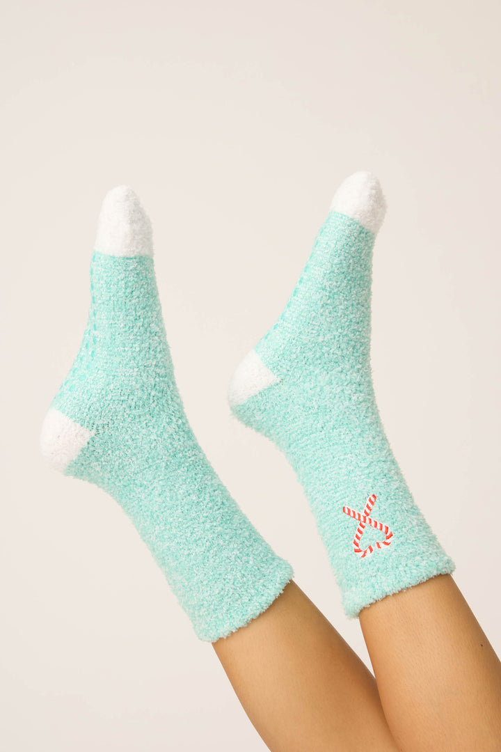 A person’s legs are raised, wearing P.J. Salvage Candy Cane Fuzzy Crew Socks in light blue with white heels and toes, featuring a small red and white candy cane on the ankle against a plain light background.