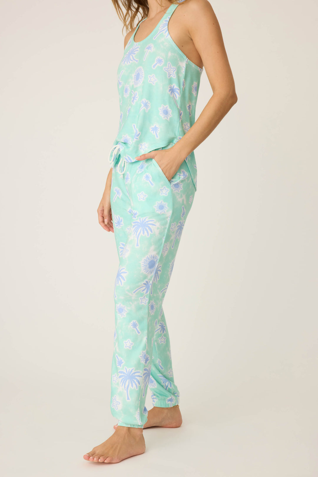 A person models the P.J. Salvage Vacay Vibes Palm Print Banded Pants, featuring a white and blue tropical pattern on light green fabric, standing barefoot with one hand in their pocket against a plain background.