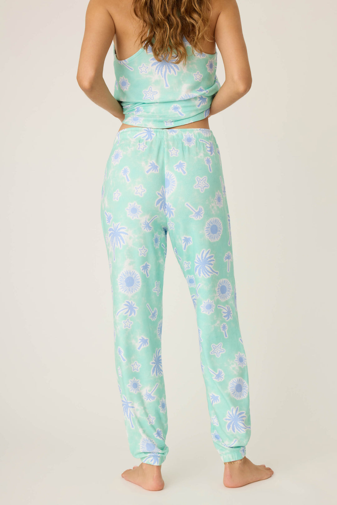 A person stands barefoot facing away, wearing P.J. Salvage's Vacay Vibes Palm Print Banded Pants in light green with a blue and white tropical pattern of palm trees, suns, and dolphins.