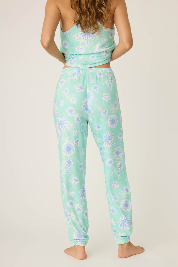 A person stands barefoot facing away, wearing P.J. Salvage's Vacay Vibes Palm Print Banded Pants in light green with a blue and white tropical pattern of palm trees, suns, and dolphins.