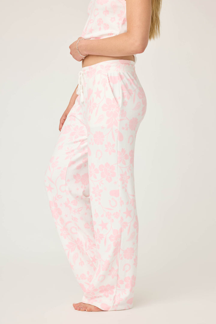 A person stands sideways wearing P.J. Salvage's Wild Flower Western Floral Print Open Leg Pants, paired with a matching top. The pajama set features white fabric with light pink floral and star patterns against a plain background.
