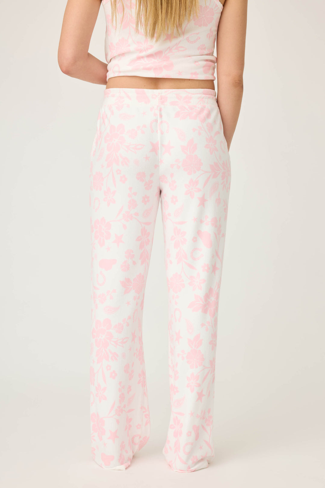 A person with long, straight hair is seen from the back wearing P.J. Salvage’s Wild Flower Western Floral Print Open Leg Pants—a relaxed-fit white pant with a light pink floral pattern—paired with a white tank top against a plain white background.