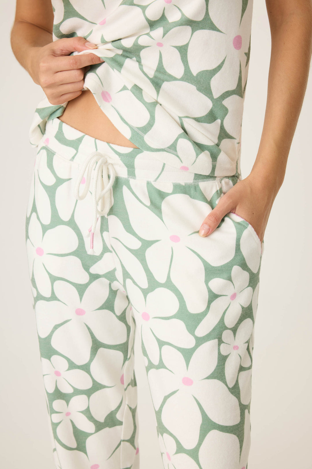 Wearing the P.J. Salvage Wish You Were Here Floral Print Banded Pants, a person shows off the green and white floral pajama set with pink accents—one hand lifts the shirt to reveal the waist, while the other rests in a pocket.